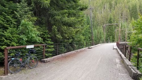 A gravel path leads over a bridge, surrounded by lush green trees, with a bicycle parked nearby.