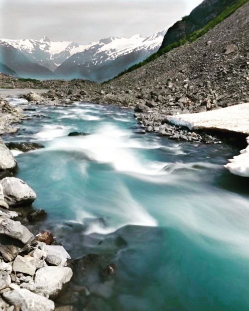 A serene river flows through rocky terrain, with snow-capped mountains in the background under a cloudy sky.