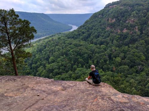 A person sits on a rocky ledge overlooking a lush green valley and winding river below.