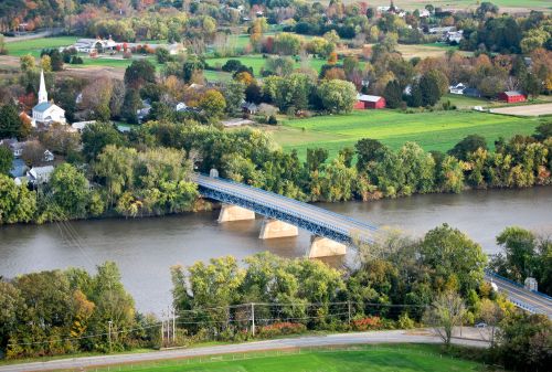 Aerial view of a river with a blue bridge, surrounded by green fields and a small town with a church steeple.