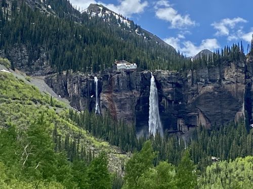 A scenic view of a waterfall cascading down a rocky cliff, surrounded by lush green trees and mountains under a blue sky.
