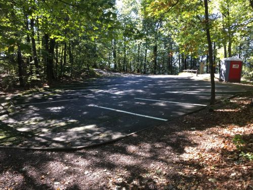 A nearly empty parking lot surrounded by trees, with a red restroom facility in the background.