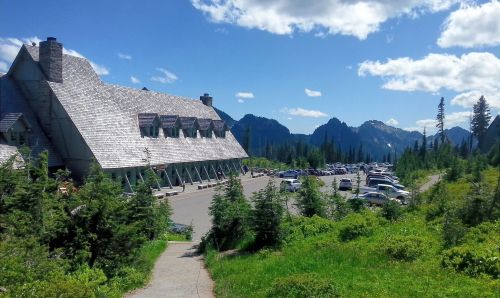 A lodge surrounded by trees and mountains, with a parking lot filled with cars under a blue sky with fluffy clouds.