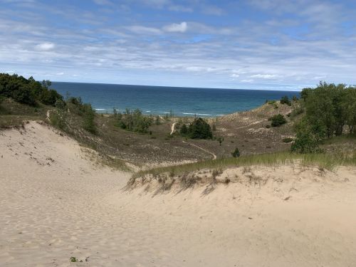 A sandy dune landscape with green vegetation, leading to a blue ocean under a partly cloudy sky.