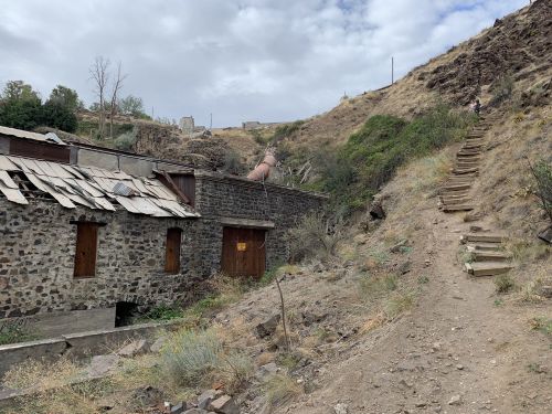 A rocky hillside with a dirt path leading up, flanked by an old stone building and overgrown vegetation.