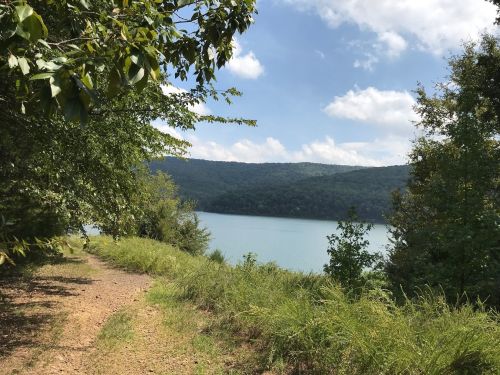 A serene lakeside path surrounded by greenery and hills under a partly cloudy sky.