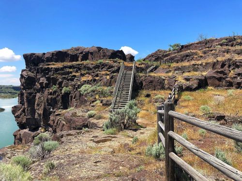 A rocky cliff with a wooden fence and stairs leading up, overlooking a calm body of water under a blue sky.