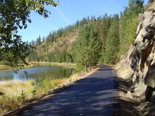 A scenic pathway alongside a river, surrounded by tall trees and rocky hills under a clear blue sky.