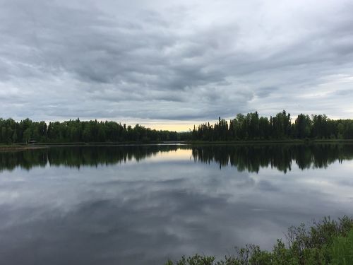 A calm lake reflects a cloudy sky and surrounding green trees, creating a serene natural landscape.