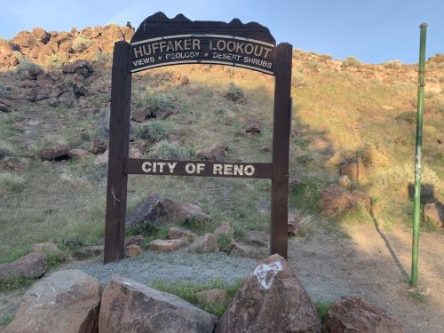 Sign for Huffaker Lookout, featuring views of geology and desert shrubs, with "City of Reno" below. Rocky hillside in background.