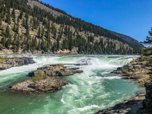 A vibrant river cascades over rocky formations, surrounded by lush green trees and mountains under a clear blue sky.
