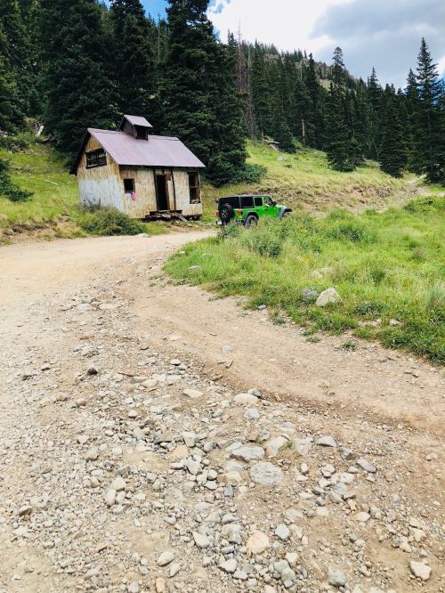 A rustic cabin beside a gravel road, with a green vehicle parked nearby, surrounded by tall trees and grassy terrain.