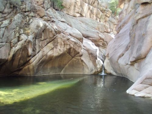 A serene pool surrounded by large, smooth rock formations and a small waterfall in a natural setting.