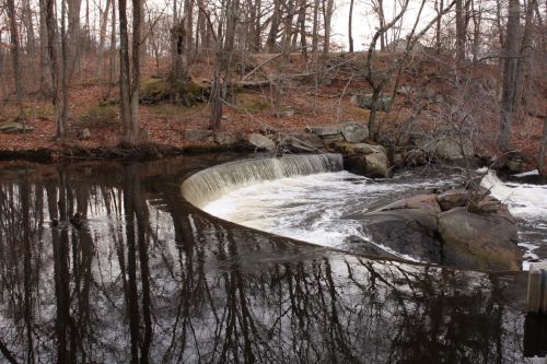 A serene scene of a curved waterfall flowing into a calm river, surrounded by bare trees and rocky terrain.