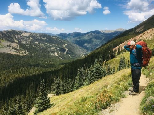 A hiker with a backpack stands on a trail, gazing at a scenic mountain view with lush greenery and blue skies.