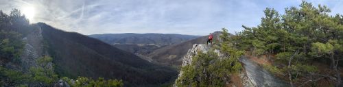 A panoramic view of a hiker on a rocky outcrop, surrounded by mountains and trees under a clear sky.