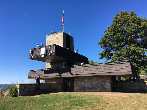 A modern stone and wood building with people on the roof, set against a clear blue sky and green trees.