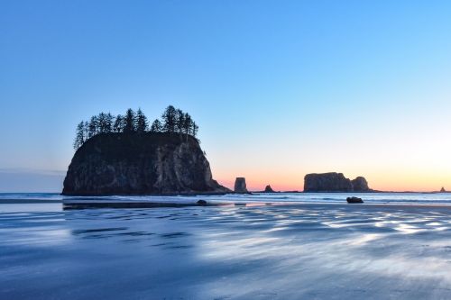 A serene beach at sunset, featuring rocky islands and a silhouette of trees on one island against a blue sky.
