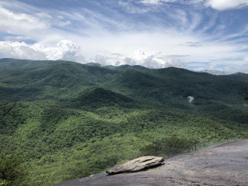 Lush green mountains stretch under a cloudy sky, with a rocky foreground and mist rising in the valleys.