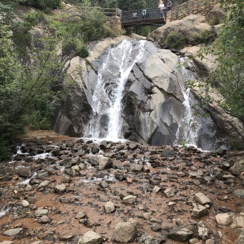 A waterfall cascades over large rocks, surrounded by greenery, with a bridge visible in the background.