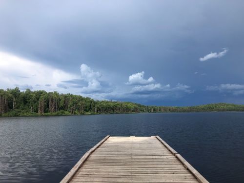 A wooden dock extends into a calm lake, surrounded by lush green trees under a cloudy sky.