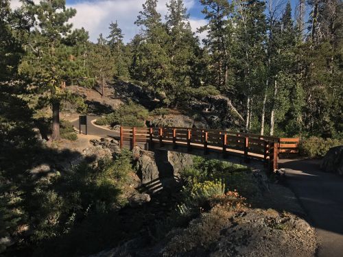 A wooden bridge spans a rocky path surrounded by trees and greenery in a serene outdoor setting.