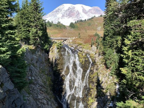 A waterfall cascades down rocky terrain, surrounded by lush greenery, with a snow-capped mountain in the background.