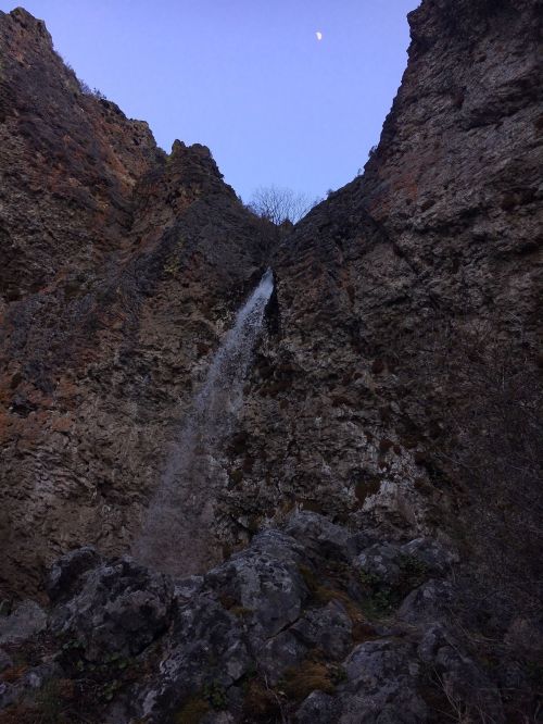 A waterfall cascades down rocky cliffs under a twilight sky with a crescent moon visible above.