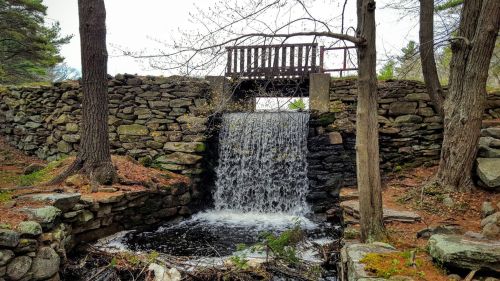 A serene waterfall flows beneath a wooden bridge, surrounded by stone walls and trees in a peaceful natural setting.