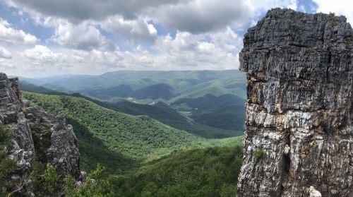 A panoramic view of lush green mountains under a cloudy sky, with a rocky cliff in the foreground.