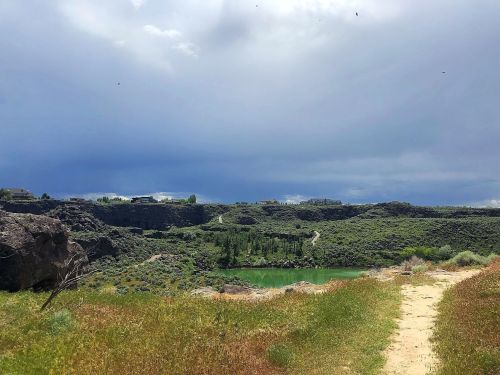A scenic view of a green lake surrounded by rocky terrain and lush grass under a cloudy sky.