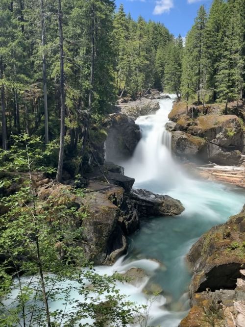 A serene waterfall cascades through lush green trees and rocky terrain under a clear blue sky.
