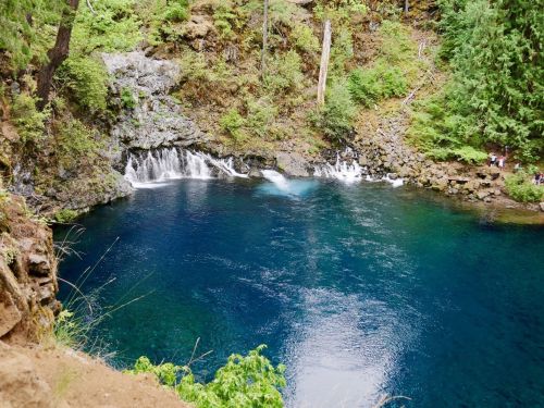 A serene blue pool surrounded by rocky cliffs and lush greenery, with a small waterfall cascading into the water.