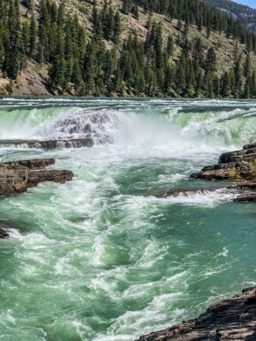 A rushing river flows over rocks, surrounded by lush green trees and steep hills under a clear blue sky.
