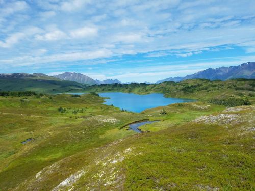 A serene landscape featuring a blue lake surrounded by green hills and mountains under a partly cloudy sky.