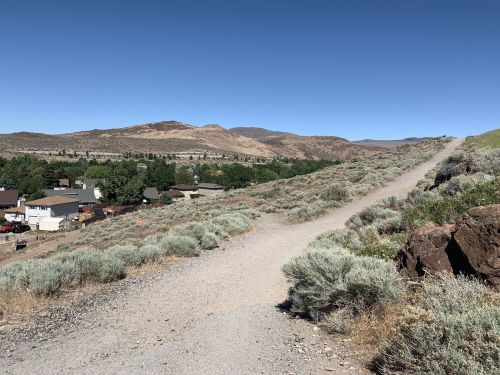 A gravel path winds through sagebrush, leading to a hillside town with mountains in the background under a clear blue sky.