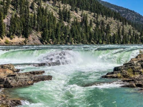 A rushing river cascades over rocky cliffs, surrounded by lush green trees and mountains in the background.