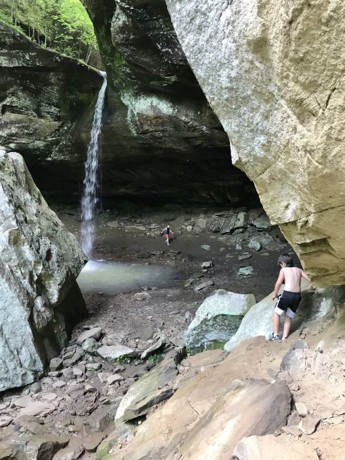A waterfall cascades into a rocky cave, with two people exploring the area below. Lush greenery surrounds the scene.