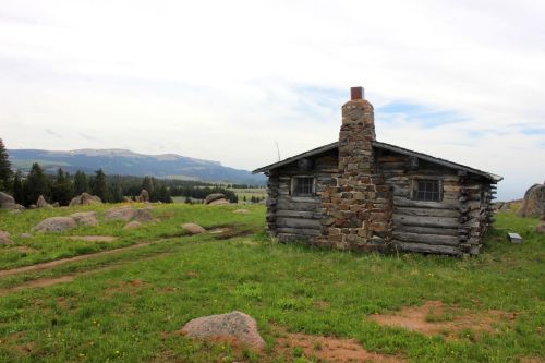 A rustic log cabin with a stone chimney sits in a grassy field, surrounded by boulders and distant mountains.