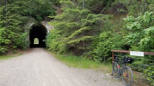 A gravel path leads to a dark tunnel, with a bike parked nearby and lush greenery surrounding the area.