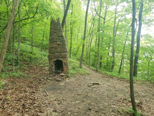 A stone chimney stands in a lush green forest, beside a winding dirt path surrounded by trees.