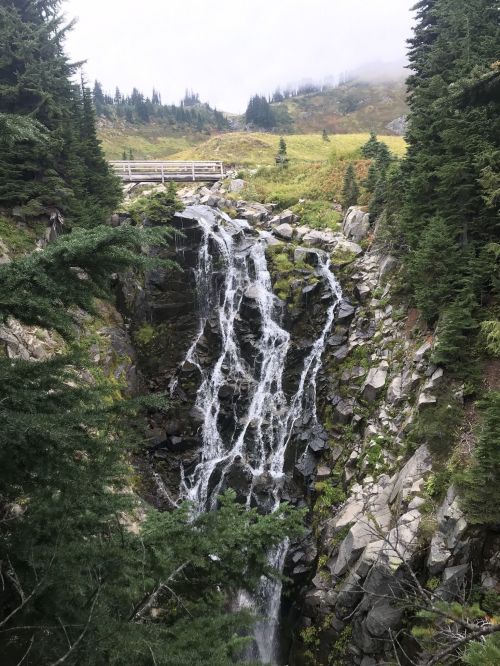 A cascading waterfall flows over rocky terrain, surrounded by lush greenery and misty hills in the background.