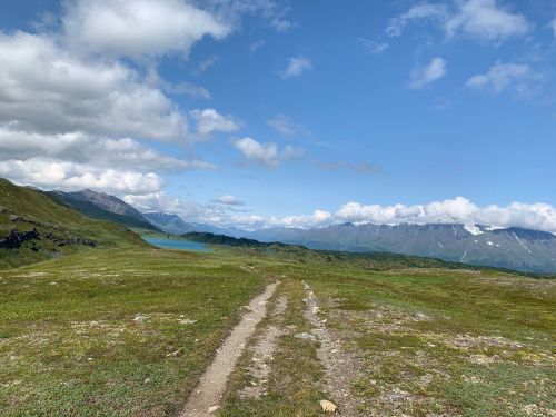 A scenic landscape featuring a dirt path leading through green hills, with mountains and a lake under a partly cloudy sky.