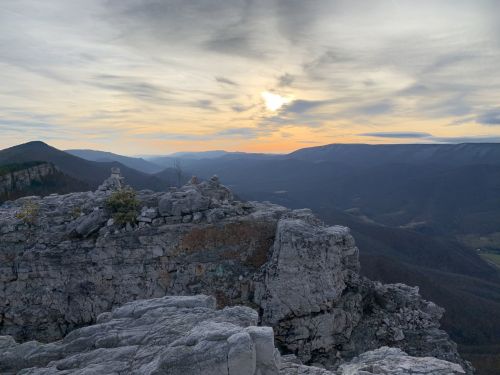 A rocky outcrop overlooks a valley at sunset, with mountains and a cloudy sky in the background.