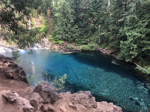 A serene blue lake surrounded by lush green trees and rocky terrain, with gentle water flowing nearby.