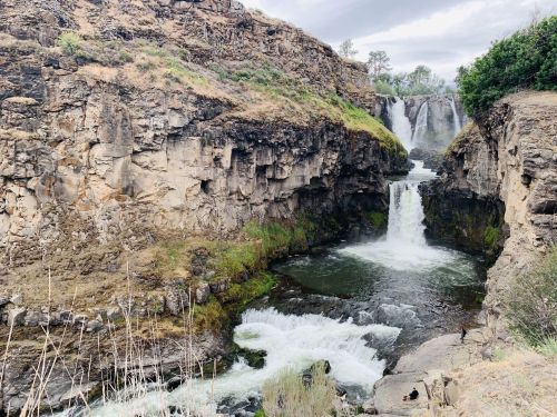 A scenic view of a waterfall cascading between rocky cliffs, surrounded by greenery and flowing water below.