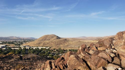 A rocky foreground overlooks a valley with a small town and rolling hills under a clear blue sky.