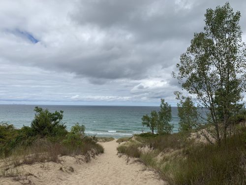 A sandy path leads to a calm beach with gentle waves under a cloudy sky, framed by green trees.