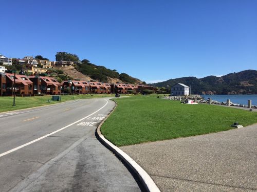 A scenic view of a road alongside a grassy area and waterfront, with houses on a hillside and mountains in the background.