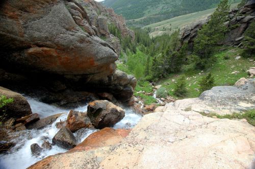 A rocky landscape with a flowing stream, surrounded by lush greenery and steep hills in the background.